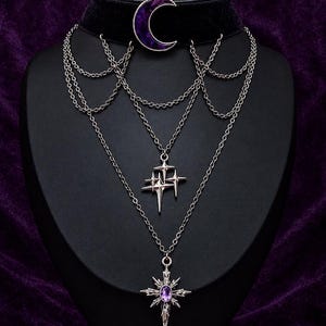May include: A black velvet choker necklace featuring a purple crescent moon charm, layered silver-tone chains, a cluster of four-pointed star charms, and a central star-shaped pendant with a small purple oval gemstone in the center.