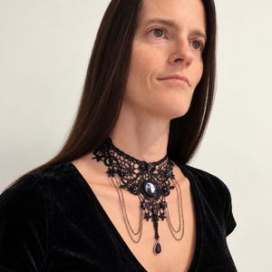 Op de afbeelding: Een zwarte kanten choker-halsketting met ingewikkelde bloemmotieven, donkerpaarse ovale cabochonstenen en delicate hangende metalen kettinkjes. Het ontwerp bevat een centrale druppelvormige paarse steen als hanger.