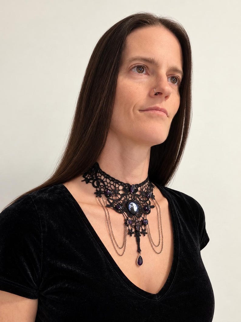 Op de afbeelding: Een zwarte kanten choker-halsketting met ingewikkelde bloemmotieven, donkerpaarse ovale cabochonstenen en delicate hangende metalen kettinkjes. Het ontwerp bevat een centrale druppelvormige paarse steen als hanger.