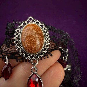 May include: A black lace choker necklace featuring a central oval goldstone cabochon set in a silver-toned filigree frame. A faceted red teardrop-shaped pendant hangs from the bottom of the frame, with additional small red beads attached to the lace.