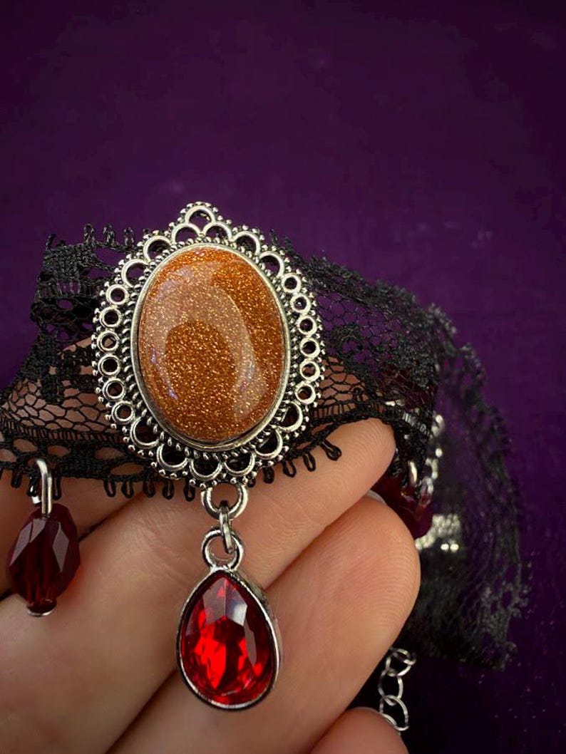 May include: A black lace choker necklace featuring a central oval goldstone cabochon set in a silver-toned filigree frame. A faceted red teardrop-shaped pendant hangs from the bottom of the frame, with additional small red beads attached to the lace.
