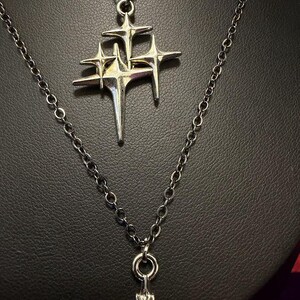 May include: A silver-toned necklace featuring a pendant shaped like a cluster of three four-pointed stars with elongated, sharp rays. The pendant hangs from a delicate silver-colored metal chain with circular links, showcasing a metallic finish.