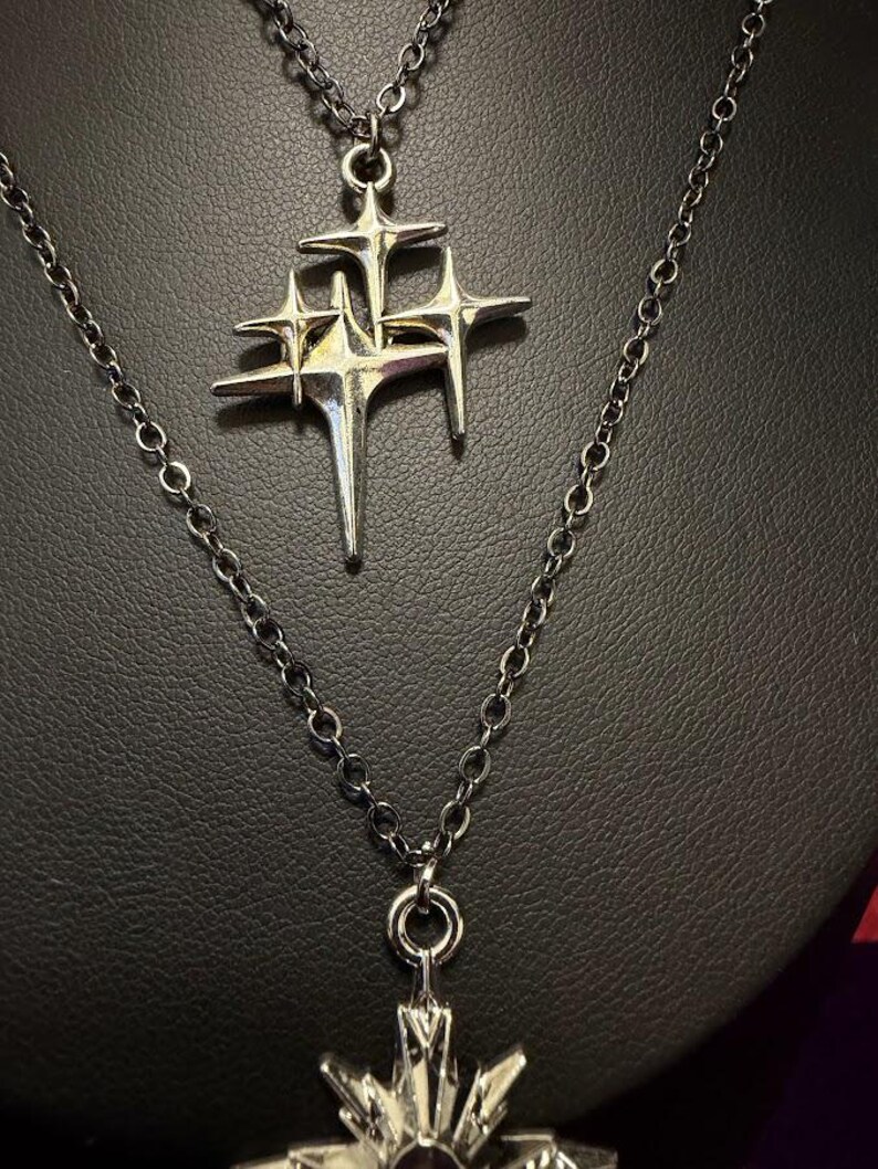 May include: A silver-toned necklace featuring a pendant shaped like a cluster of three four-pointed stars with elongated, sharp rays. The pendant hangs from a delicate silver-colored metal chain with circular links, showcasing a metallic finish.
