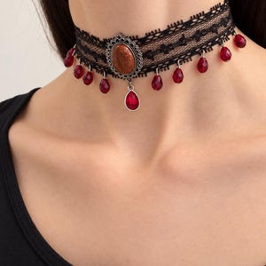May include: A black lace choker necklace featuring a central oval-shaped brown glittery cabochon in a silver-toned filigree setting, accented with multiple dangling red teardrop-shaped beads suspended along the lower edge of the lace band.