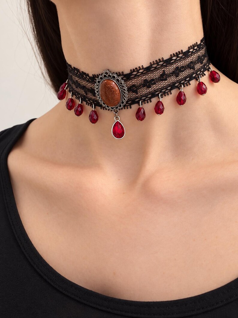 May include: A black lace choker necklace featuring a central oval-shaped brown glittery cabochon in a silver-toned filigree setting, accented with multiple dangling red teardrop-shaped beads suspended along the lower edge of the lace band.