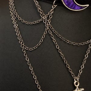 May include: A black velvet choker necklace featuring a purple crescent moon pendant with a silver-toned rim. The piece includes multiple delicate silver-toned metal chains that drape downwards, connecting to a silver-toned star-shaped charm at the base.