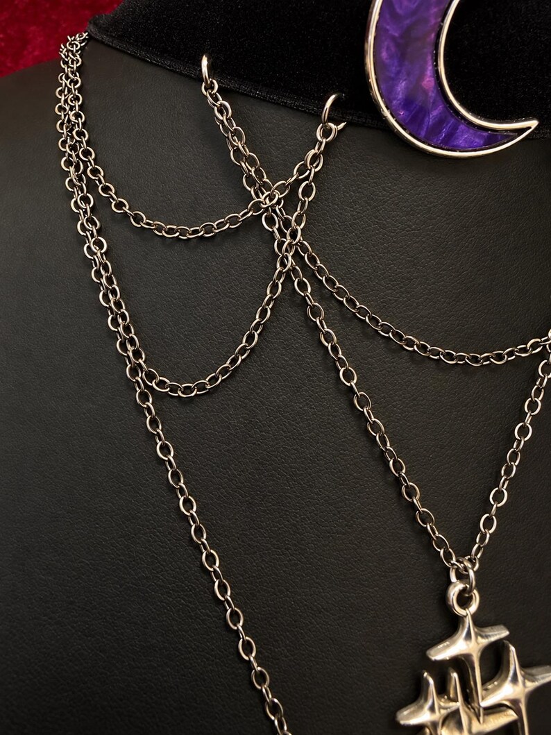 May include: A black velvet choker necklace featuring a purple crescent moon pendant with a silver-toned rim. The piece includes multiple delicate silver-toned metal chains that drape downwards, connecting to a silver-toned star-shaped charm at the base.
