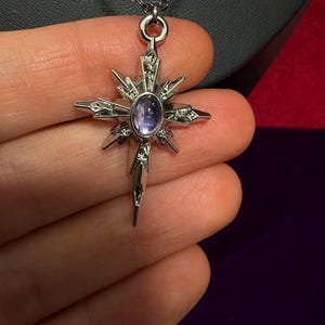 May include: A silver-toned metal pendant shaped like a multi-pointed star or compass rose, featuring a central oval-shaped purple cabochon stone. The pendant is suspended from a thin silver-toned chain and has intricate etched detailing on its rays.