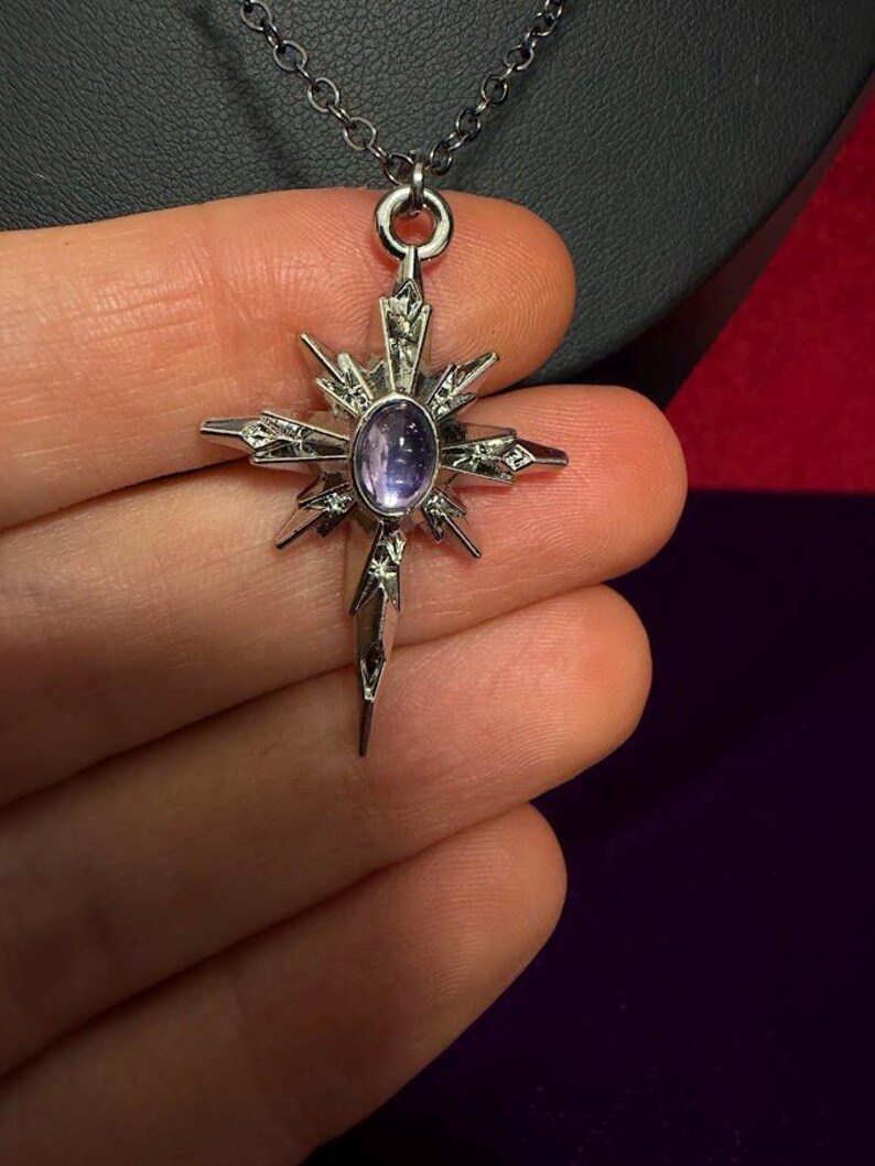 May include: A silver-toned metal pendant shaped like a multi-pointed star or compass rose, featuring a central oval-shaped purple cabochon stone. The pendant is suspended from a thin silver-toned chain and has intricate etched detailing on its rays.