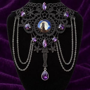 Op de afbeelding: Een zwarte kanten choker met een centraal rond cabochon dat het silhouet van een kat tegen een volle maan toont. Het sieraad is versierd met meerdere gefacetteerde paarse kristallen en fijne zilverkleurige metalen kettinkjes.