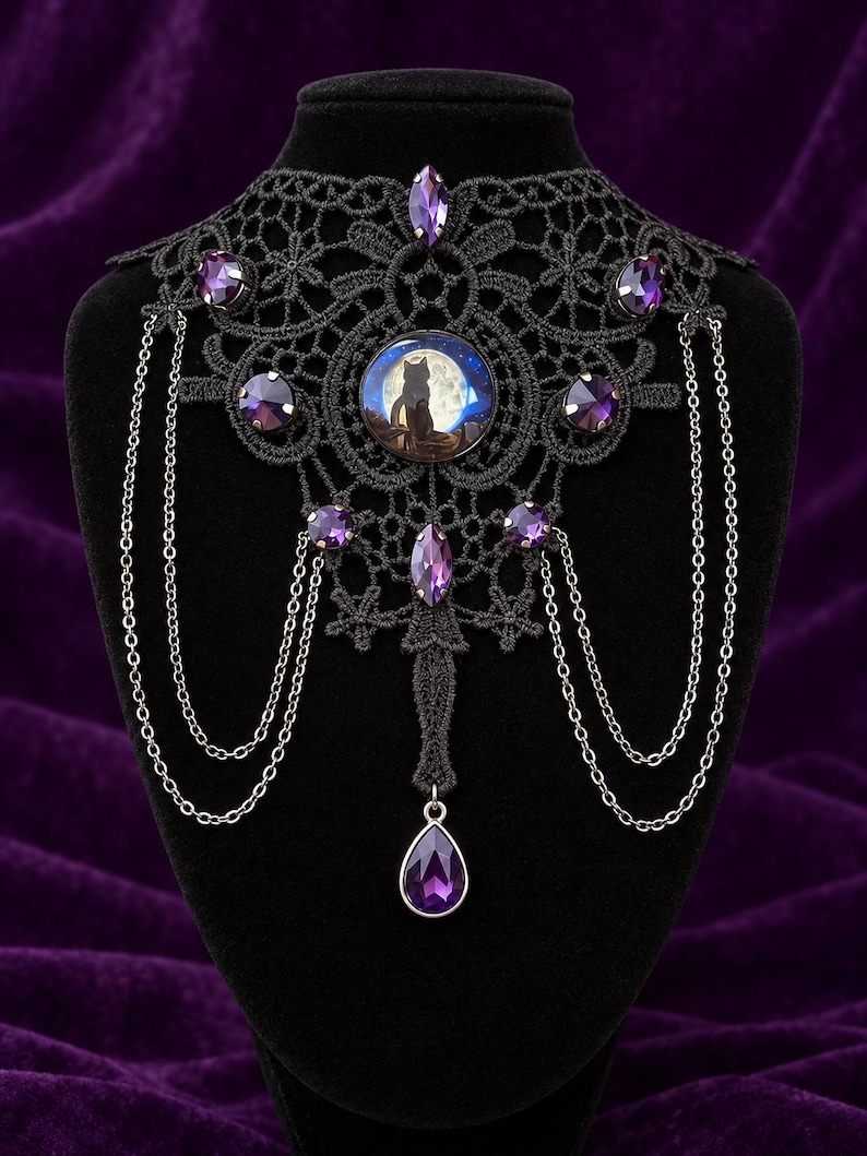 Op de afbeelding: Een zwarte kanten choker met een centraal rond cabochon dat het silhouet van een kat tegen een volle maan toont. Het sieraad is versierd met meerdere gefacetteerde paarse kristallen en fijne zilverkleurige metalen kettinkjes.