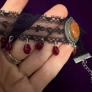 May include: A black lace choker necklace featuring a central oval-shaped orange glittery cabochon set in a silver-toned metal frame, accented with multiple dangling dark red faceted teardrop beads and a silver-toned metal chain extension.