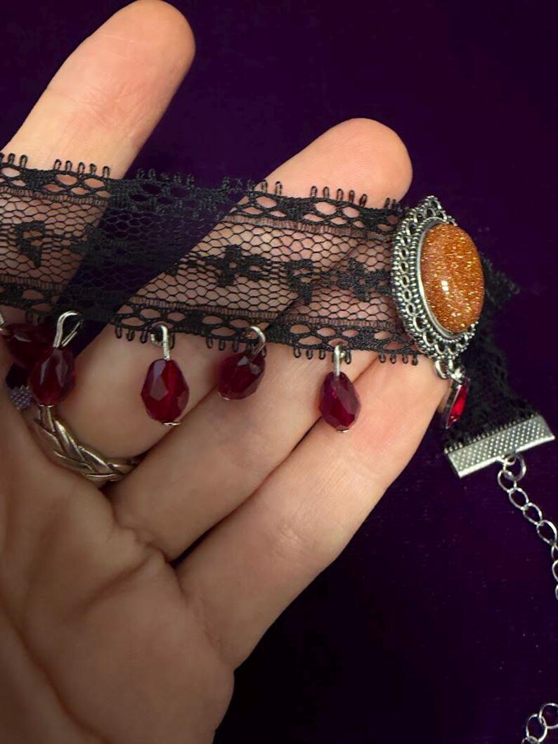 May include: A black lace choker necklace featuring a central oval-shaped orange glittery cabochon set in a silver-toned metal frame, accented with multiple dangling dark red faceted teardrop beads and a silver-toned metal chain extension.