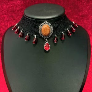 May include: A black lace choker necklace featuring a central oval-shaped goldstone cabochon set in a silver-toned filigree frame. A single red teardrop-shaped crystal hangs from the center, with smaller red crystal beads dangling along the lace.
