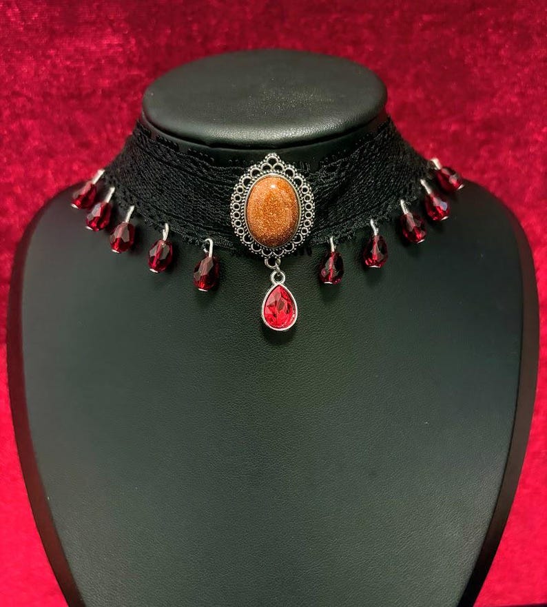 May include: A black lace choker necklace featuring a central oval-shaped goldstone cabochon set in a silver-toned filigree frame. A single red teardrop-shaped crystal hangs from the center, with smaller red crystal beads dangling along the lace.
