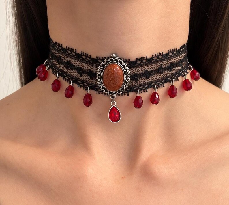 May include: A black lace choker necklace featuring a central oval-shaped brown glittery stone set in a silver-toned filigree frame. The choker is adorned with multiple small red teardrop-shaped beads hanging from the bottom edge of the lace band.
