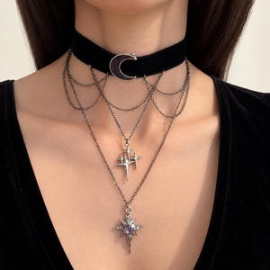 May include: A black velvet choker necklace featuring a silver-toned crescent moon charm. The piece includes multiple draped silver chains connecting to two lower pendants: one shaped like a cluster of four-pointed stars and one starburst charm.