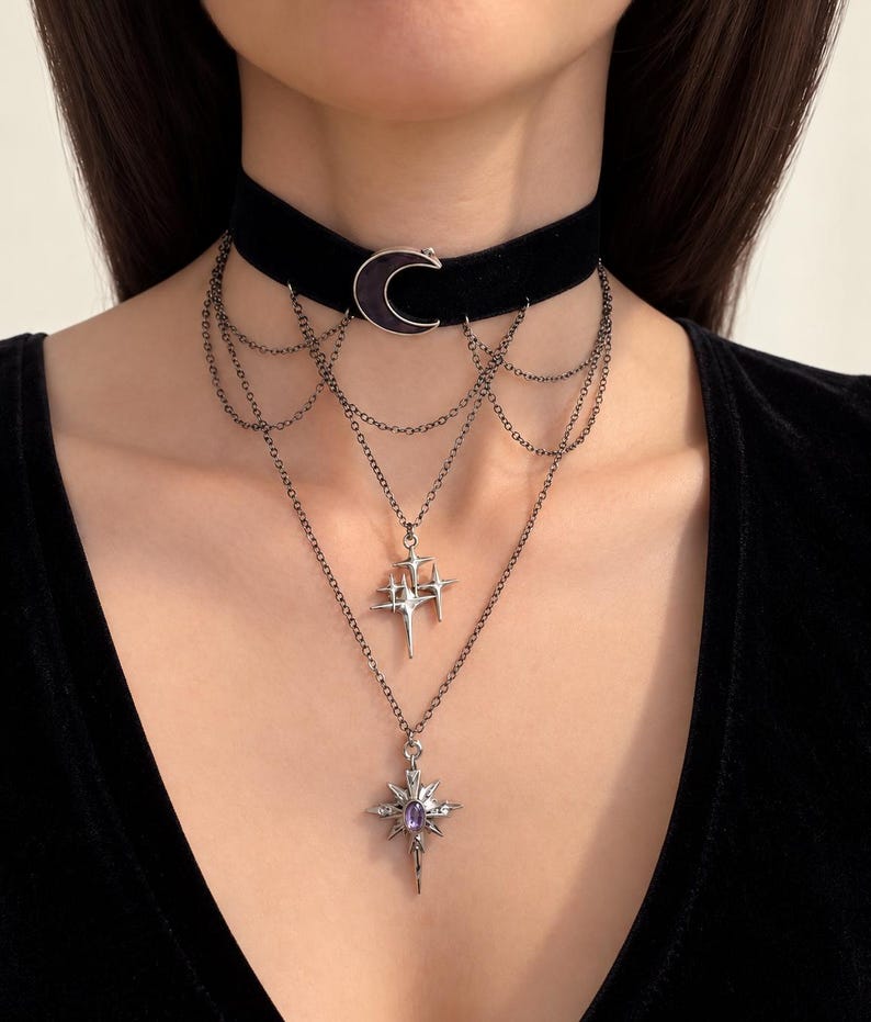 May include: A black velvet choker necklace featuring a silver-toned crescent moon charm. The piece includes multiple draped silver chains connecting to two lower pendants: one shaped like a cluster of four-pointed stars and one starburst charm.