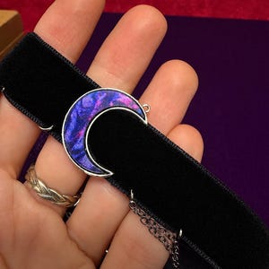 May include: A black velvet choker necklace featuring a silver-toned crescent moon pendant. The moon pendant is filled with a swirling, iridescent pattern of deep purple, blue, and magenta colors. A thin silver-toned metal chain hangs from the side.