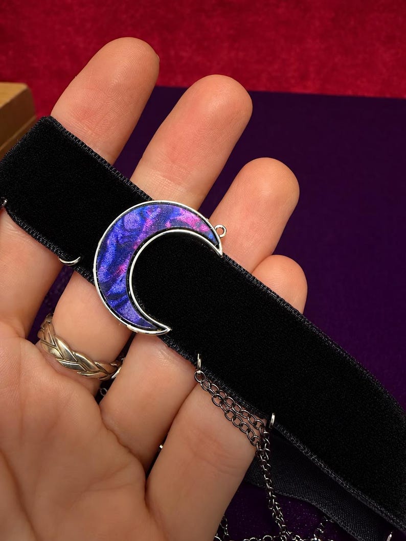 May include: A black velvet choker necklace featuring a silver-toned crescent moon pendant. The moon pendant is filled with a swirling, iridescent pattern of deep purple, blue, and magenta colors. A thin silver-toned metal chain hangs from the side.