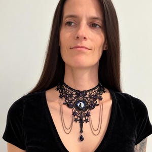 Op de afbeelding: Een zwarte kanten choker-halsketting met een centrale ronde cabochon met een silhouet van een kat, geaccentueerd met dieppaarse gefacetteerde kristallen, fijne hangende metalen kettinkjes en een druppelvormige paarse kristallen hanger.