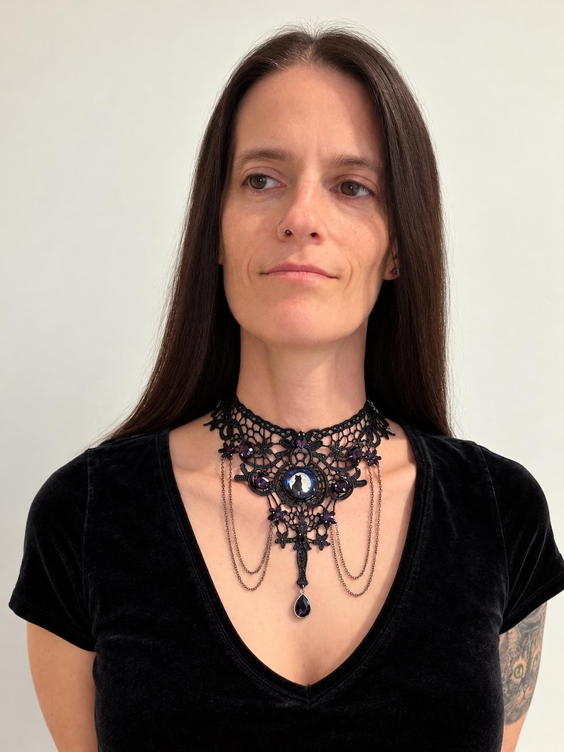 Op de afbeelding: Een zwarte kanten choker-halsketting met een centrale ronde cabochon met een silhouet van een kat, geaccentueerd met dieppaarse gefacetteerde kristallen, fijne hangende metalen kettinkjes en een druppelvormige paarse kristallen hanger.