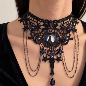 Op de afbeelding: Een zwarte kanten chokerhalsketting met een centrale ronde cabochon met het silhouet van een kat tegen een volle maan. Het ontwerp bevat meerdere paarse gefacetteerde kristallen, fijne zwarte metalen kettinkjes en een druppelvormige hanger.