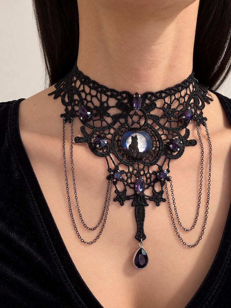 Op de afbeelding: Een zwarte kanten chokerhalsketting met een centrale ronde cabochon met het silhouet van een kat tegen een volle maan. Het ontwerp bevat meerdere paarse gefacetteerde kristallen, fijne zwarte metalen kettinkjes en een druppelvormige hanger.