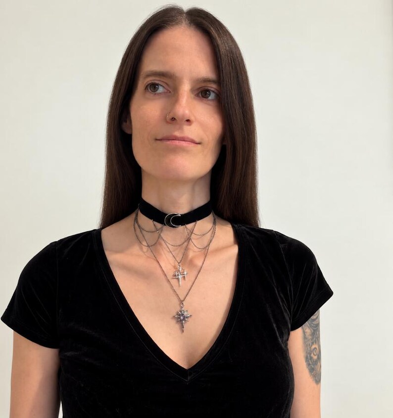 May include: A black velvet choker necklace featuring a silver-toned crescent moon charm at the center. Attached to the choker are multiple delicate silver-toned chains that drape downwards, ending in two distinct star-shaped pendants with small crystals.