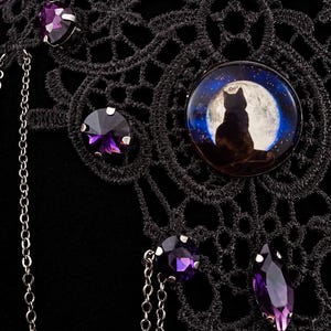 Op de afbeelding: Een zwarte kanten choker met een ronde cabochon waarop het silhouet van een kat voor een volle maan is afgebeeld. Het sieraad is versierd met meerdere gefacetteerde paarse kristallen in zilverkleurige zettingen en fijne metalen kettinkjes.