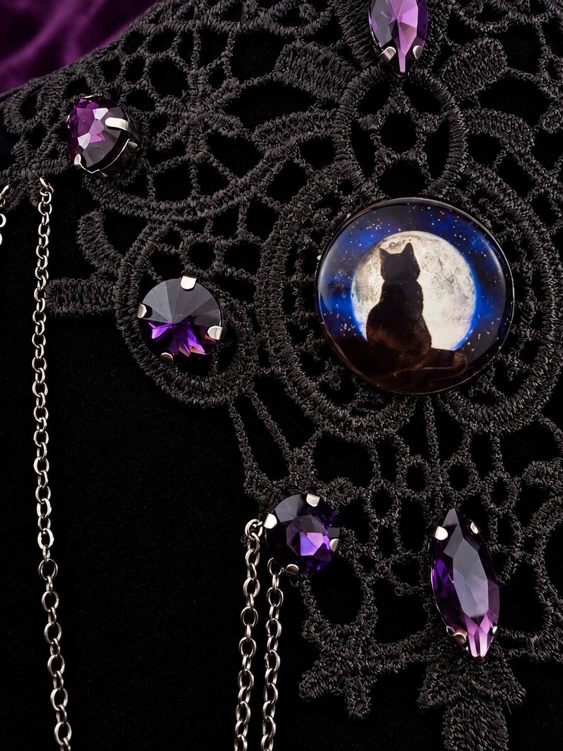Op de afbeelding: Een zwarte kanten choker met een ronde cabochon waarop het silhouet van een kat voor een volle maan is afgebeeld. Het sieraad is versierd met meerdere gefacetteerde paarse kristallen in zilverkleurige zettingen en fijne metalen kettinkjes.