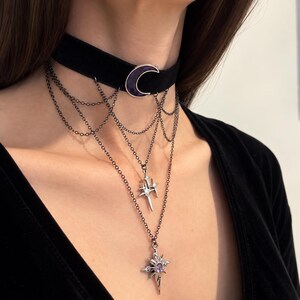 May include: A black velvet choker necklace featuring a central purple crescent moon charm. The piece includes multiple draped dark metal chains connected to two silver-toned star-shaped pendants, with the lower pendant set with a small purple stone.