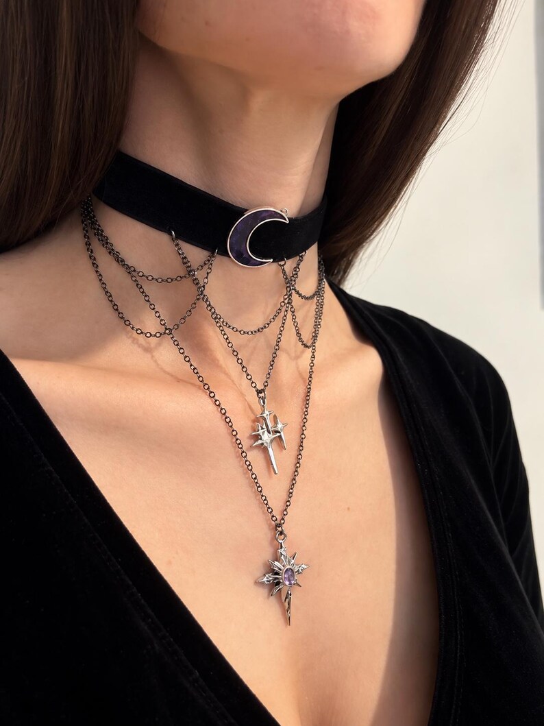 May include: A black velvet choker necklace featuring a central purple crescent moon charm. The piece includes multiple draped dark metal chains connected to two silver-toned star-shaped pendants, with the lower pendant set with a small purple stone.