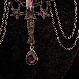 Op de afbeelding: Een zwarte kanten choker-halsketting met een centrale paarse kristal in marquise-slijpvorm, twee ronde paarse kristallen accenten en een bungelende druppelvormige paarse kristallen hanger. Het ontwerp bevat fijne zilverkleurige metalen kettingen.