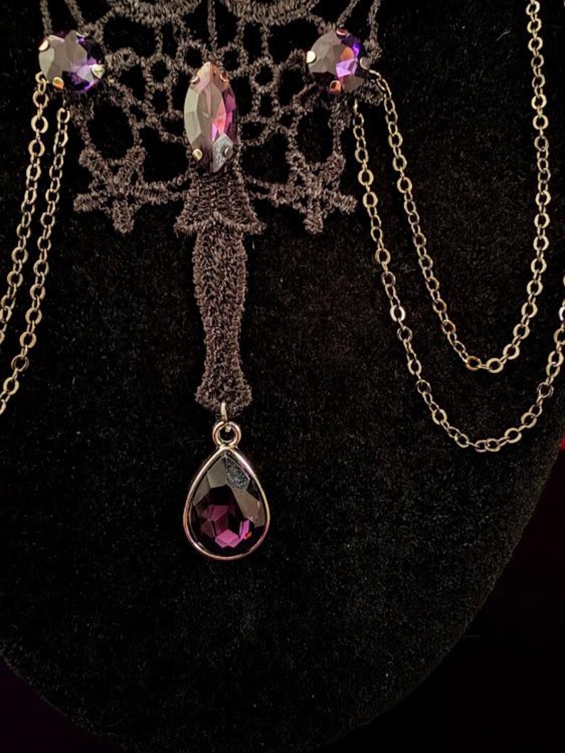 Op de afbeelding: Een zwarte kanten choker-halsketting met een centrale paarse kristal in marquise-slijpvorm, twee ronde paarse kristallen accenten en een bungelende druppelvormige paarse kristallen hanger. Het ontwerp bevat fijne zilverkleurige metalen kettingen.