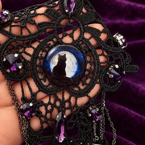 Op de afbeelding: Een zwarte kanten choker met een centrale ronde cabochon met het silhouet van een kat tegen een volle maan. Het kant is versierd met meerdere gefacetteerde paarse kristallen in zilverkleurige zettingen en bevat fijne hangende zilveren kettinkjes.