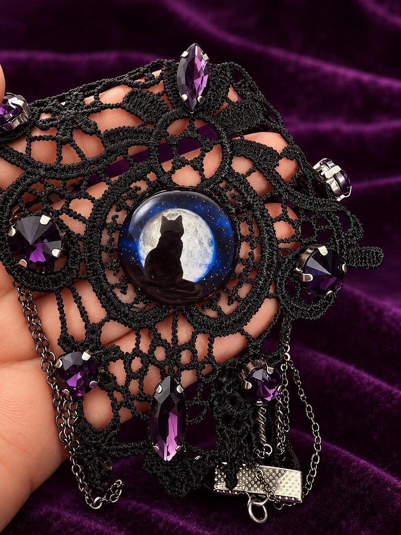 Op de afbeelding: Een zwarte kanten choker met een centrale ronde cabochon met het silhouet van een kat tegen een volle maan. Het kant is versierd met meerdere gefacetteerde paarse kristallen in zilverkleurige zettingen en bevat fijne hangende zilveren kettinkjes.