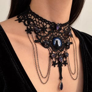 Op de afbeelding: Een zwarte kanten choker ketting met een centrale ovale cabochon met het silhouet van een kat tegen een volle maan. Het sieraad is versierd met donkerpaarse gefacetteerde kristallen en fijne hangende metalen kettinkjes met een druppelhanger.