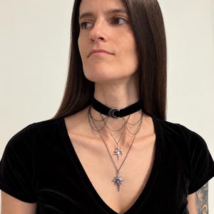 May include: A black velvet choker necklace featuring a silver-toned crescent moon charm at the center. The piece includes multiple layered silver-toned chains that drape downwards, adorned with a small star charm and a larger star-shaped pendant.