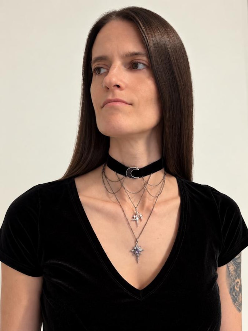 May include: A black velvet choker necklace featuring a silver-toned crescent moon charm at the center. The piece includes multiple layered silver-toned chains that drape downwards, adorned with a small star charm and a larger star-shaped pendant.