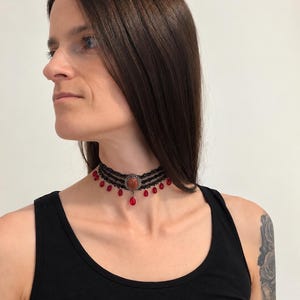 May include: A black lace choker necklace featuring three horizontal bands of intricate lace trim. The center is adorned with a round, copper-toned setting holding a smooth, reddish-brown stone. Small, teardrop-shaped red beads hang from the lace.