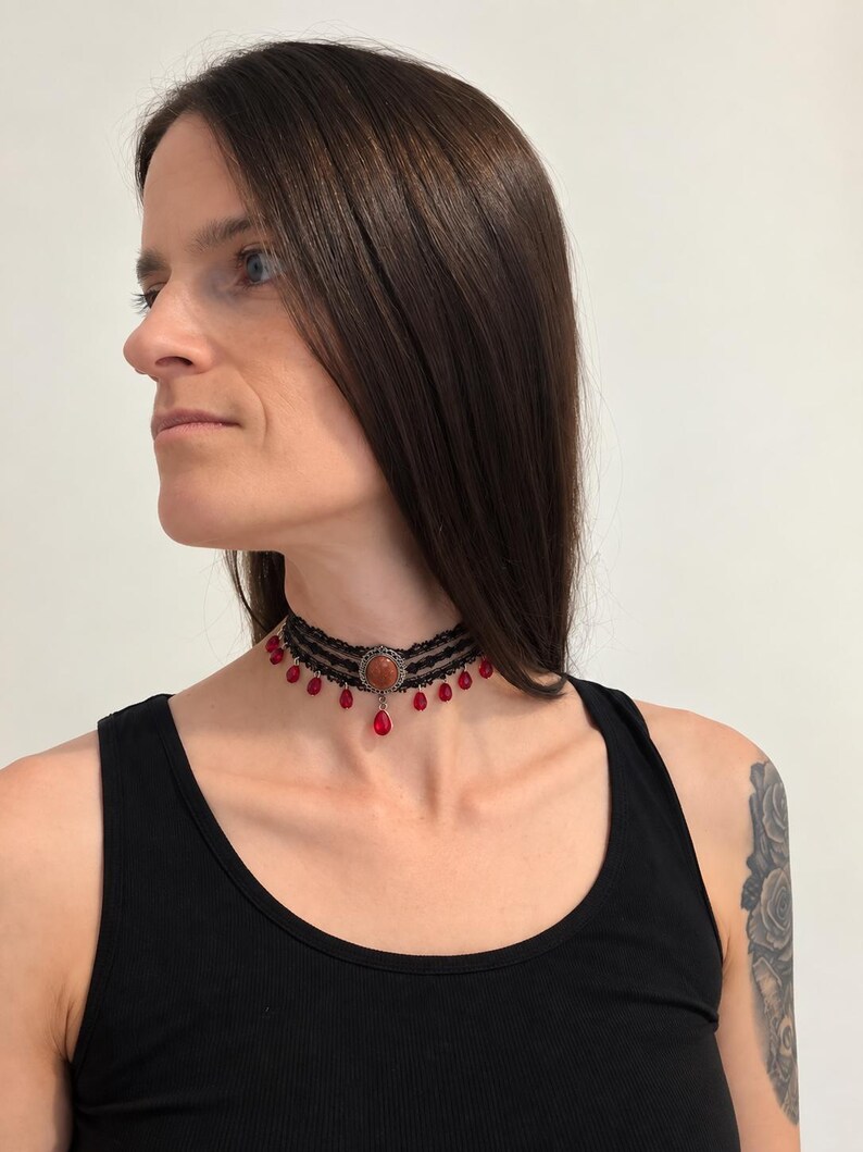 May include: A black lace choker necklace featuring three horizontal bands of intricate lace trim. The center is adorned with a round, copper-toned setting holding a smooth, reddish-brown stone. Small, teardrop-shaped red beads hang from the lace.