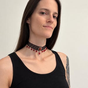 May include: A black lace choker necklace featuring a central oval-shaped amber-colored stone set in a silver-toned frame, accented with multiple dangling red teardrop-shaped beads hanging from the bottom edge of the lace band.