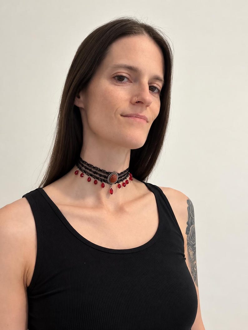 May include: A black lace choker necklace featuring a central oval-shaped amber-colored stone set in a silver-toned frame, accented with multiple dangling red teardrop-shaped beads hanging from the bottom edge of the lace band.