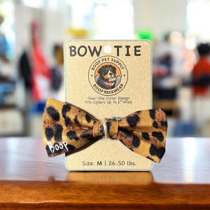 May include: A leopard print bow tie for dogs, with the word "boop" printed on the corner. The bow tie is attached to a cardboard tag that reads "BOW TIE" and "BOOP PET SUPPLY". The tag also states the bow tie fits collars up to 1" wide.