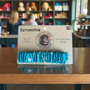 Puede incluir: Una scrunchie turquesa para perros, con un diseño floral y de lunares blancos, exhibida en un soporte de cartón. El embalaje incluye el texto "Scrunchie", "Boop Pet Supply" y "Over-the-Collar Design". Talla S, para perros de 5 a 11 kg.