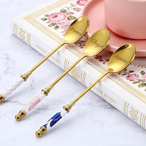 May include: Three gold-colored spoons with engraved names: Penny, Birgit, and Agnini. Each spoon has a decorative handle with white, pink, and blue enamel accents. The spoons rest on a floral-patterned book next to a pink teacup and saucer.