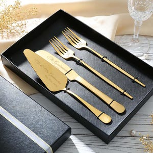 May include: A gold-colored cake server and two forks with the text "Mr. & Mrs. Groomsmans" and a date engraved on the server. The utensils are in a black box, with a gift box and champagne glass in the background.