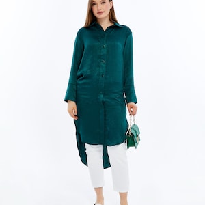 May include: An emerald green tunic shirt with long sleeves and a button-down design, worn with white cropped trousers and white loafers. A small, green handbag is held. The shirt has a high-low hemline.