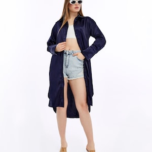 May include: A long, navy blue button-down shirt with long sleeves. The shirt is worn open over a white crop top and light blue denim shorts. The model is wearing clear heeled sandals and white sunglasses.